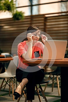 Portrait of confident mature professional woman in glasses, a coral T-shirt sitting on summer terrace in cafe, using laptop