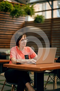 Portrait of confident mature professional woman in glasses, a coral T-shirt sitting on summer terrace in cafe, using laptop