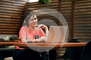 Portrait of confident mature professional woman in glasses, a coral T-shirt sitting on summer terrace in cafe, using laptop