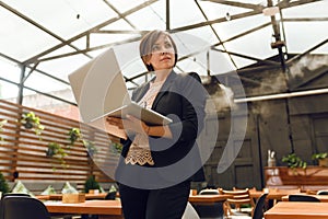 Portrait of confident mature professional woman in glasses, a coral T-shirt sitting on summer terrace in cafe, using laptop