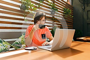 Portrait of confident mature professional woman in glasses, a coral T-shirt sitting on summer terrace in cafe, using laptop