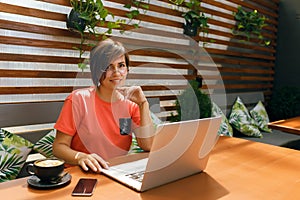 Portrait of confident mature professional woman in glasses, a coral T-shirt sitting on summer terrace in cafe, using laptop