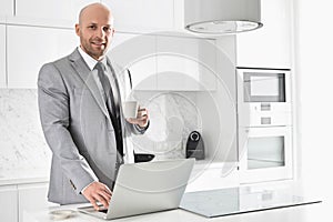 Portrait of confident mid adult businessman having coffee while using laptop in kitchen