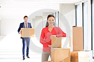 Portrait of confident mid adult businesswoman standing by stacked boxes with male colleague in background at office