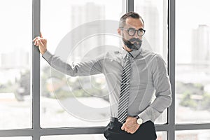 Portrait of confident middle-aged businessman in blue shirt and wearing glasses with a beard and mustache standing next to window