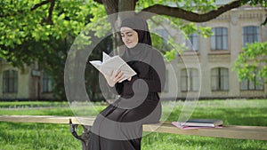 Portrait of confident Muslim woman enjoying reading outdoors on sunny day. Happy young university student sitting at