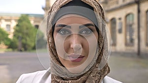 Portrait of confident muslim woman in hijab looking at camera, standing on street near university building