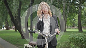 Portrait of confident positive businesswoman talking on smartphone standing in summer park with bicycle. Beautiful