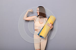 Portrait of confident prod woman wearing beige top and leggins holding fitness mat  over gray background, raised her arm,