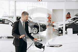 Portrait of a confident sales manager in the suit near the car charging station, selling electric cars in the showroom