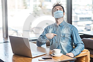 Portrait of confident satisfied egoistic young man with surgical medical mask in blue shirt sitting, working on laptop and