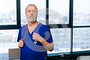 Portrait of confident senior adult male doctor in medical uniform standing in hospital office on background of window