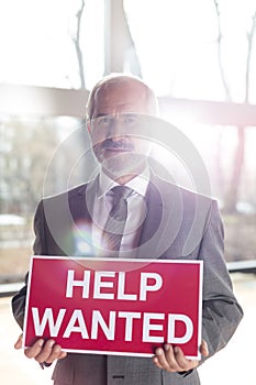 Portrait of confident senior businessman holding help wanted sign while standing at office