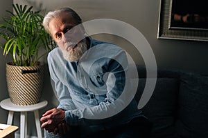 Portrait of confident senior gray-haired male sitting on sofa by bedside lamp in dark living room and looking camera.