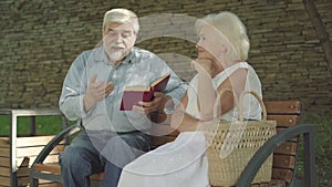 Portrait of confident senior man reading book in park for beautiful Caucasian woman. Happy husband and wife sitting on