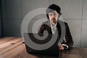 Portrait of confident serious businessman in suit sitting at workplace with laptop looking at camera. Successful CEO