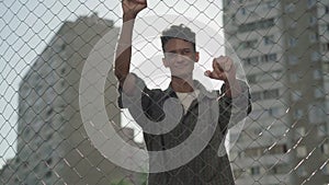 Portrait of confident smiling mixed-race man posing in sunrays outdoors. Happy young handsome guy holding mesh fence and