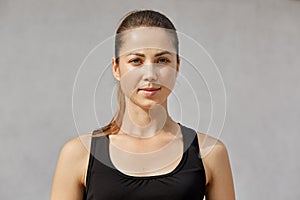 Portrait of confident sporty woman with ponytail standing over gray background and looking at camera, has serious facial