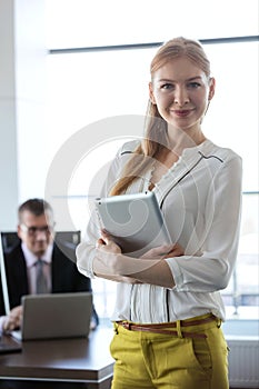 Portrait of confident young businesswoman holding tablet PC with businessman in background at office