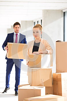 Portrait of confident young businesswoman standing by stacked boxes with male colleague in background at office