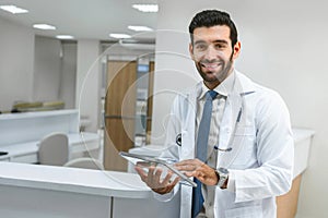 Portrait of confident young doctor holding transparent tablet and standing smiling in hospital