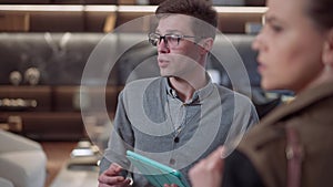Portrait of confident young male seller in eyeglasses with tablet standing in furniture shop talking to client. Expert