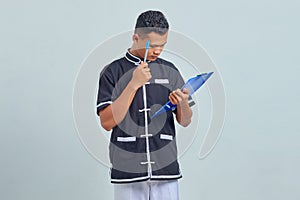 Portrait of confused asian young man wearing karate uniform holding clipboard and pen with pen