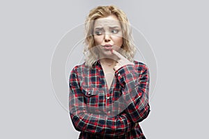 Portrait of confused beautiful blonde young woman in casual red checkered shirt standing, looking away and thinking what to do