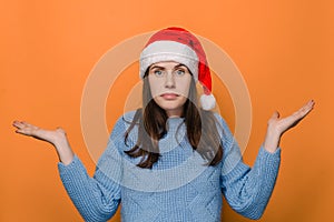 Portrait of confused beautiful woman in Christmas hat, spreads hands sideways, smirks face, feels doubt while makes choice