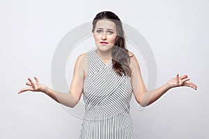 Portrait of confused beautiful young brunette woman with makeup and striped dress standing and looking at camera with crossed arms