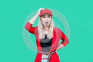 Portrait of confused blond young hipster woman in red blouse, cap, standing with hand on waist and on head looking away shocked,