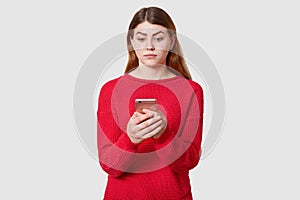 Portrait of confused cute woman with dark straight hair, wears red sweater, holds mobile phone in hand, looking at its display,