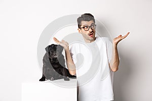 Portrait of confused hipster guy dog owner shrugging, standing near cute black pug pet, white background
