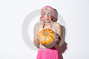 Portrait of confused and scared girl with pink wig and bright makeup, holding pumpkin for halloween and looking left