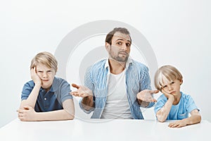 Portrait of confused unaware european father sitting with sons at table, shrugging with spread palms and questioned