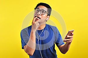 Portrait of confused Young Asian man bitting his finger and listening live music from smartphone with headset