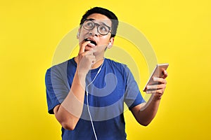 Portrait of confused Young Asian man bitting his finger and listening live music from smartphone with headset