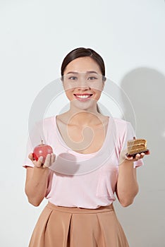 Portrait of a confused young asian woman choosing between cakes and apple, isolated on a white background