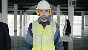 Portrait of construction specialist wearing safety uniform standing in empty building with businesspeople in background
