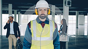 Portrait of construction specialist wearing safety uniform standing in empty building with businesspeople in background