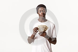 Portrait of cool african-american guy in beanie counting money, standing white background
