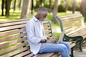Portrait of cool black guy using laptop on bench at park, working remotely, communicating on webcam