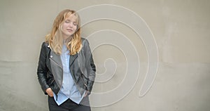 Portrait of cool blonde girl in leather jacket with hands in her pockets watching sidewards dreamily on gray wall