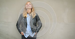 Portrait of cool blonde girl in leather jacket leaning on gray wall watching sidewards dreamily outdoors.