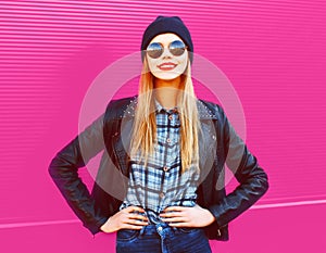 Portrait cool blonde smiling girl in rock black style jacket, hat posing on city street over colorful pink wall