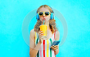 Portrait cool girl drinking fruit juice holding phone listening to music in wireless headphones on colorful blue