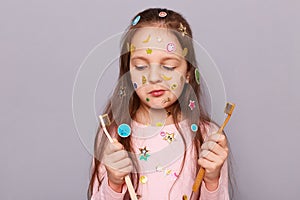 Portrait of cool little girl covered with colorful stickers wearing pink shirt, choosing toothbrush, dental care, standing