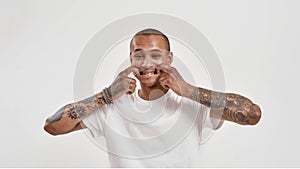 Portrait of cool mixed race tattooed guy looking at camera, forcing smile with fingers in his mouth while posing