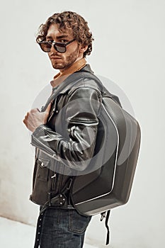 Portrait of a handsome guy with curly hair posing in the bright studio wearing sunglasses, leather coat and a backpack