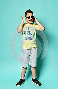 Portrait of a cool young teen guy talking on a mobile phone in summer style clothes in sunglasses, on a blue background.